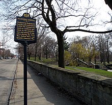 The low stone wall is seen running along the 40th Street side of Arsenal Park with the Pennsylvania Historical Marker for the Allegheny Arsenal to the left and, on the right behind some trees, a portion of the low-lying stone powder magazine is visible in the park.