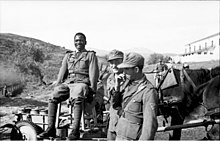 An African in German uniform sitting on a chair, next to two other soldiers having a cigarette