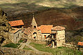 Vue en contre-bas d'un petit monastère composé de bâtiments disparates (tourelle, constructions en maçonnerie) en pierre de taille au centre desquels domine un clocher surmonté d'une croix.