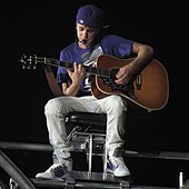 Justin Bieber playing the guitar and singing wearing a blue shirt and white pants.