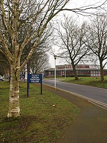 Building with multiple glass windows seen at the end of a tree lined road. Sign says welcome to Norton Radstock College