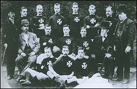 Fifteen men posing across three rows. Eleven of the men are wearing a football kit with a Maltese Cross on the breast. The other four are wearing suits and top hats.