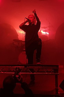 Will Ramos standing onstage, singing into a microphone and raising his other hand, with a red backlight