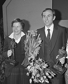 Black and white photo of Annie Borckink wearing a skirt and jacket and Joop Zoetemelk wearing a suit and tie, while both holding a statuette and flowers
