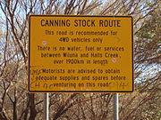 Yellow road sign in a wooded area. The title of the sign reads "Canning Stock Route". The body of the sign reads "This road is recommended for 4WD vehicles only. There is no water, fuel or services between Wiluna and Halls Creek, over 1900km in length. Motorists are advised to obtain adequate supplies and spares before venturing on this road"