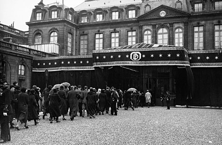 Photo en noir et blanc d’une foule d’individus faisant la queue pour accéder à un bâtiment officiel