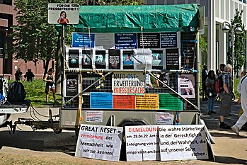 A booth mounted on a car trailer, with various signs containing beliefs about a Great Reset, including one that says (translated from German): "The same puppets who think nuclear war is justifiable, on the other hand, tell you a flu virus with 0.05 percent mortality will kill us all."