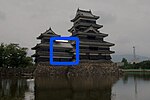 A 2-storied structure connecting a 3-storied castle tower with a 5-stoired tower. All three structures have black wooden walls and are located on a platform of unhewn stones above a water filled moat.