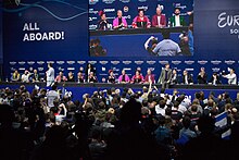 A press conference during the 2012 contest; the Serbian delegation are seated at a long table with rows of journalists seated facing them, with a large screen on the wall behind the delegation projecting a live relay of the conference.