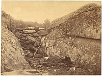 Black and white photograph of a dead soldier, lying on his back, facing slightly towards the lens, amidst rocks.