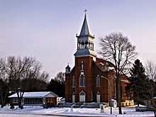 Photographie hivernale du village de Saint-Armand sous la neige. En arrière-plan se trouve l'église du village, aux briques rougeâtres et disposant d'un clocher.