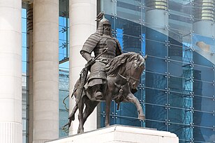 Statue of an armoured man, in front of a large pillared building.