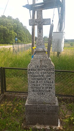 An Orthodox votive cross with Cyrillic inscriptions from 1960, located at the entrance to the village from the provincial road. In the background, there is a bus stop in the direction of Hajnówka