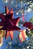 An out-of-focus close-up of a part of a Christmas tree with glitter garlands and blurry decoration. In-focus in the left half of the picture, suspended from one of the twigs, is a red cardboard-woven Froebel star. Four tips and seven prongs are visible.