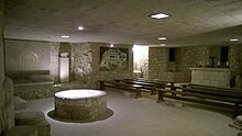 Photo d'une salle en pierre, sur la droite des bancs et un autel avec une croix.