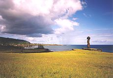 Several Hanga Roa moai, including Ko Te Riku (with a pukao on its head). In the mid-ground is a side view of an ahu with five moai. The Mataveri end of Hanga Roa is visible in the background with Rano Kau rising above it.