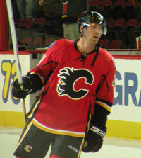 Hockey player in red uniform, with a "C" in the middle. He leans to his left, holding his stick by his side.