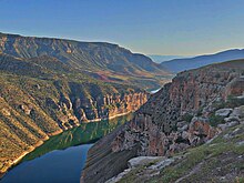 Grand canyon alternant falaises et pentes couvertes d'une végétation éparse. La roche est rouge-orangé, et la végétation verte mais sèche. L'eau reflète le bleu du ciel.