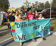 Three children are seen holding a banner which says "Different NOT Less! We ARE UNIFIED" in brightly colored text.