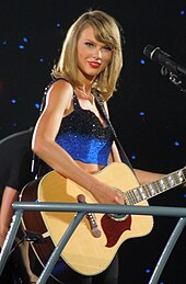 Taylor Swift in a black and blue crop top, holding a brown acoustic guitar
