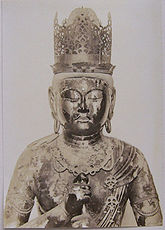 Front view, head and shoulder portrait of a statue depicted wearing a crown and showing the Vajra Mudrā, enclosing the index finger of the left hand with the fist of the right hand. Black and white picture.