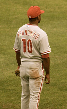 A view from the back of a dark-skinned young man in a gray baseball uniform and red baseball cap; the rear of his jersey reads "Francisco" in block red letters and "10" in larger red numbers