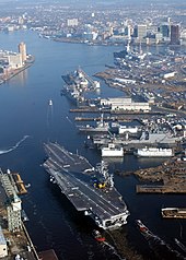 A large gray aircraft carrier sails down a river where the shore is lined with piers and large buildings.