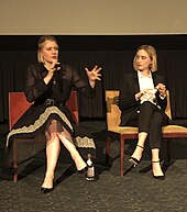 Photographie de deux femmes habillées en noir et assises devant un écran de projection. Celle de gauche, habillée en robe, tient un micro et est en train de parler, tandis que celle de droite la regarde.