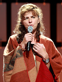 A photograph of Mira Furlan, a woman who is about 60 years old and has brown hair. She is wearing a red shawl and is singing in this image.