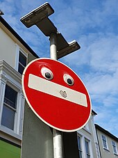Medium-sized googly eyes attached to the upper half of a red circular sign with a thick white horizontal line signifying "no entry" in front of a row of buildings