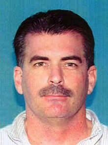 A portrait photo mugshot of a white man with dark hair; he is wearing a white collared shirt, and looking straight into the camera with a neutral expression.