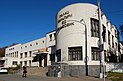 Ein recht wuchtiges, mit weißen Platten verkleidetes dreistöckiges Gebäude, zu dessen Eingang eine breite Treppe hinaufführt. Im Vordergrund dominiert ein halbrunder Gebäudeteil mit großen Fenstern. Das Gebäude liegt in der Sonne, eine Passantin geht vorüber.