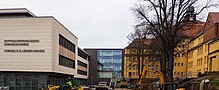 ein neues blockhaftes Gebäude auf der linken, weiß mit bunten Streifen verziert, und ein gelber Bau im Stil des frühen 20. Jahrhunderts; beide Gebäude sind im Hintergrund verbunden, die offene Seite des Hofes zeigt Richtung Kamera; auf dem Hof stehen Baumaschinen