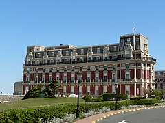 Vue d'un grand immeuble à trois étages ; façades couleur brique ou pierre et brique ; toiture en ardoises.