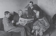 Black-and-white photograph of three men in suits and one woman seated in a room and looking at an open newspaper