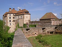 Photographie d'un bâtiment de plusieurs étages avec des toits en tuiles et construit sur un rocher qui surplombe une forêt. L'entrée principale bâtiment et la place qui se trouve devant sont entourées de murs de fortification.