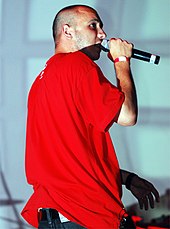 A picture of a man wearing a red shirt and jeans. He is performing in front of a grey background and holds a microphone in his right hand