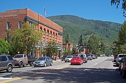 A wide undivided street with cars traveling in both directions going past a large brick building. In the rear is a forested mountain