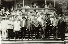 A crown of approximately 40 men standing on a set of steps in front of stone balcony