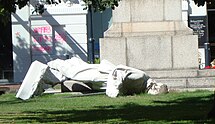 A white marble fallen statue depicting Robert Falcon Scott, on the ground in a small urban park