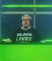 The Blessed Madonna performing on a green-lit stage wearing a black shirt that says "black lives"