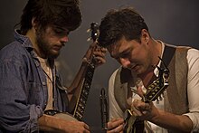 Medium shot of two men, close together, playing instruments.