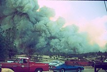 A dark, roiling gray smoke plume covers two-thirds of the sky, rising above oak woodlands with multiple parked cars in the foreground
