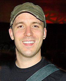 Adam Kowalczyk wearing olive green cap, black t-shirt, with strap over left shoulder and small earring in left ear, grinning at camera
