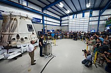 Musk is pictrued delivering a speech to SpaceX employees while standing at a podium besides a capsule in 2012
