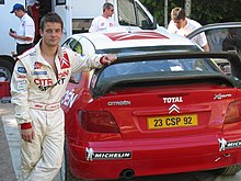 Photo de Sébastien Loeb vu de face en combinaison automobile, accoudé à l'aileron arrière d'une Citroën Xsara WRC rouge avec trois mécaniciens de l'équipe en arrière plan.