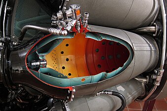 Rolls-Royce Nene with nine combustion chambers. The cutaway is one of 2 chambers fitted with a flame igniter which places the igniter in a cooler location than directly in the hot gas stream. During a start atomized fuel from the small self-contained unit (orange-coloured solenoid shown) is ignited by its ignition plug and the flaming jet of fuel is projected into the main fuel spray from the burner. Combustion is propagated to all the chambers through interconnecting tubes.[89]