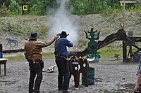 Um atirador de CAS disparando um rifle de alavanca em alvos de aço. O Range Officer à esquerda está segurando um cronômetro para medir o tempo.