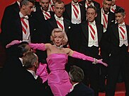 Marilyn Monroe in a pink gown, surrounded by men in tuxedos