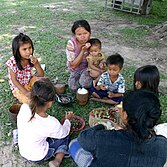 Two Southeast Asian women and five children sit on grass eating rice and vegetables